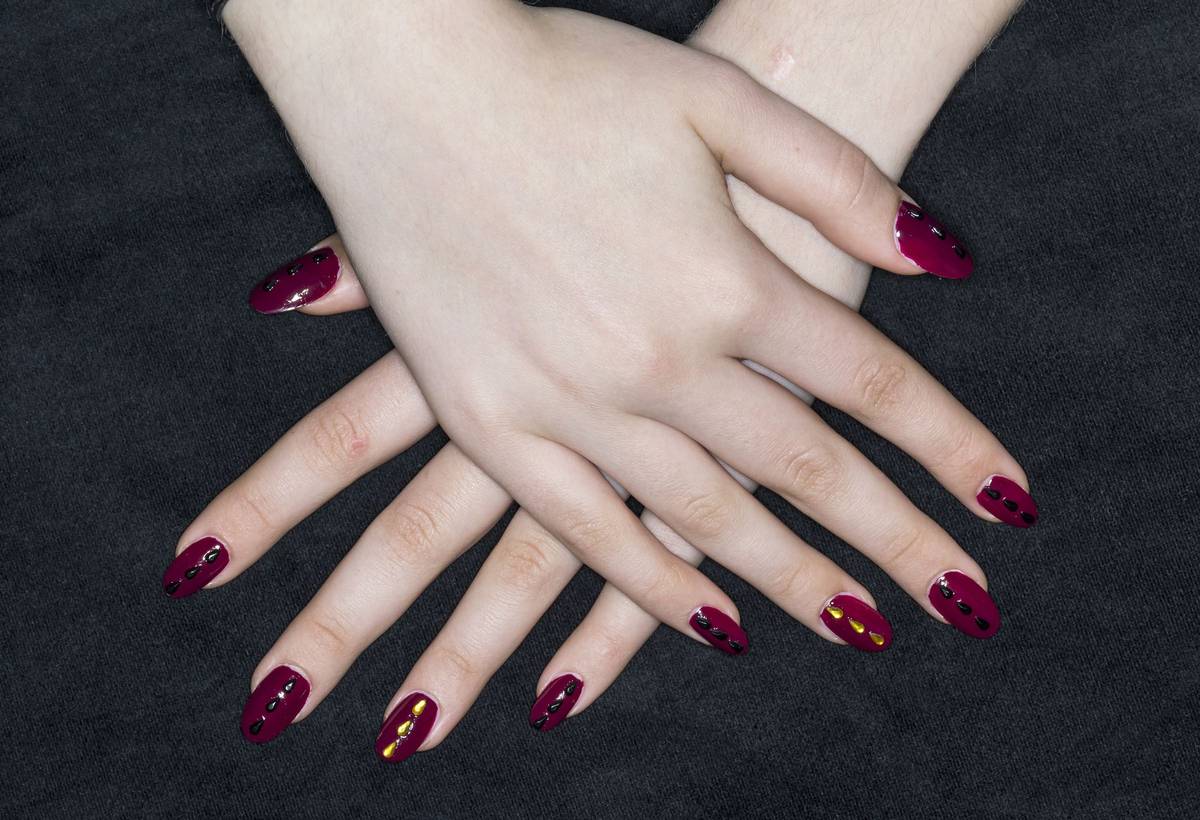 Two crossed hands on dark fabric, nails painted deep plum with small black and gold studs
