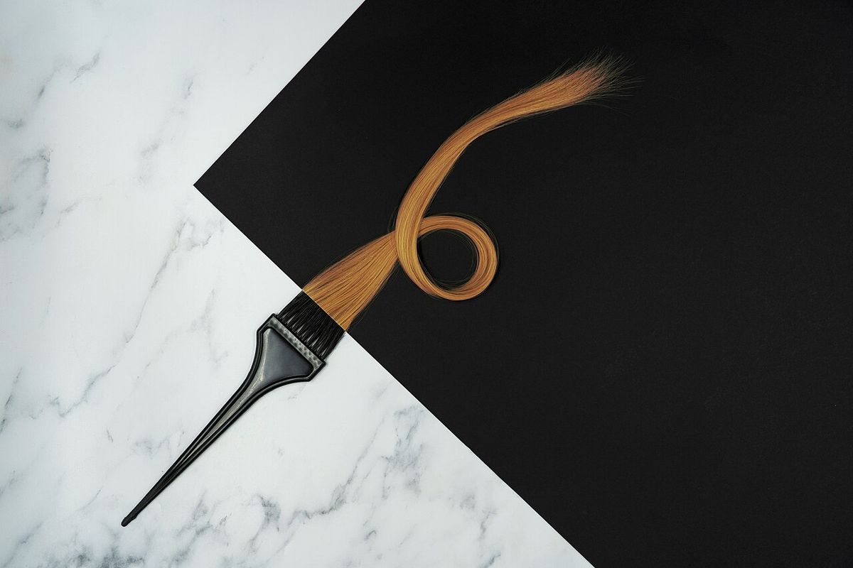 A hair-colour tint brush beside a curled lock of copper-brown hair on black paper and white marble
